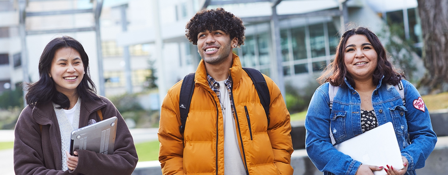Three students walking in a line on a university campus while they are smiling.