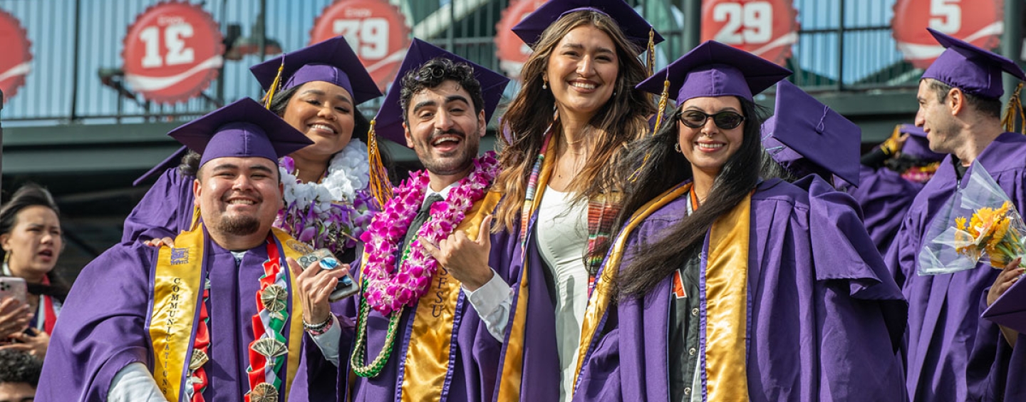 Graduates dressed in regalia stand together in the bleachers at Oracle Park