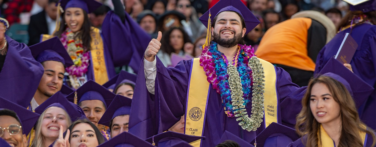 A crowd of smiling graduates dressed in reglia with one grad standing giving thumbs up