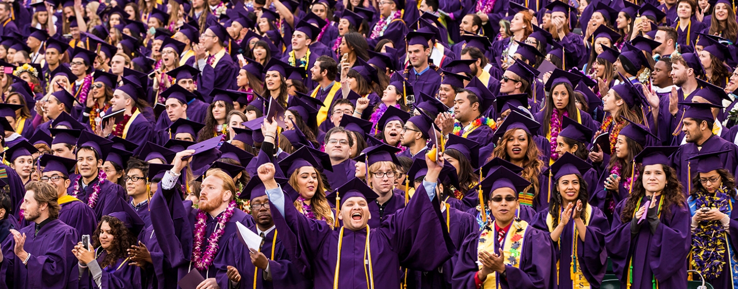 Several students stand in a stadium dressed in their caps and gown while one student smiles directly at the camera gesturing triumphantly
