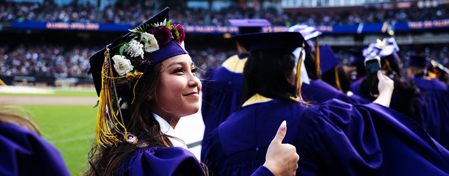 Student dressed in regalia in a ballpark gives the thumbs up