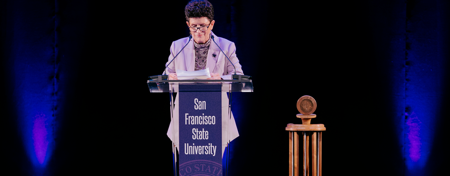 President Lynn Mahoney speaks behind a lectern decorated in purple with the text San Francisco State University in front of a black background and next to a wooden gavel 