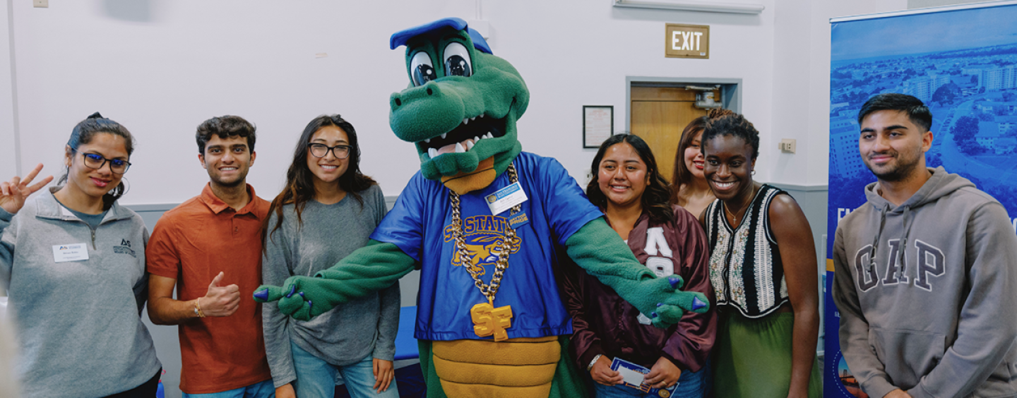 Seven people smile while posing for a photo with the Alli Gator mascot in the middle pointing their fingers horizontally and with one of the other people flashing a peace sign