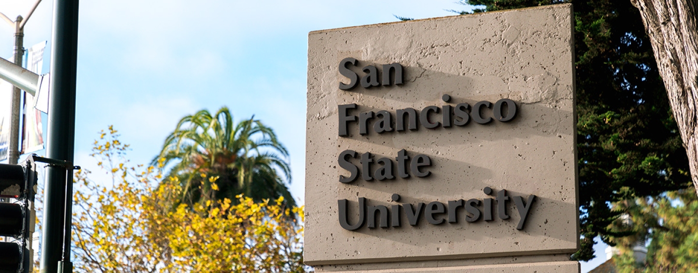 The San Francisco State University pylon sign surrounded by trees and next to a stoplight and a bicyclist road sign on a mostly sunny day