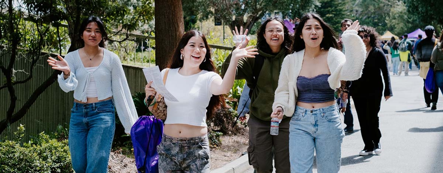 Four women waving to camera while walking on campus