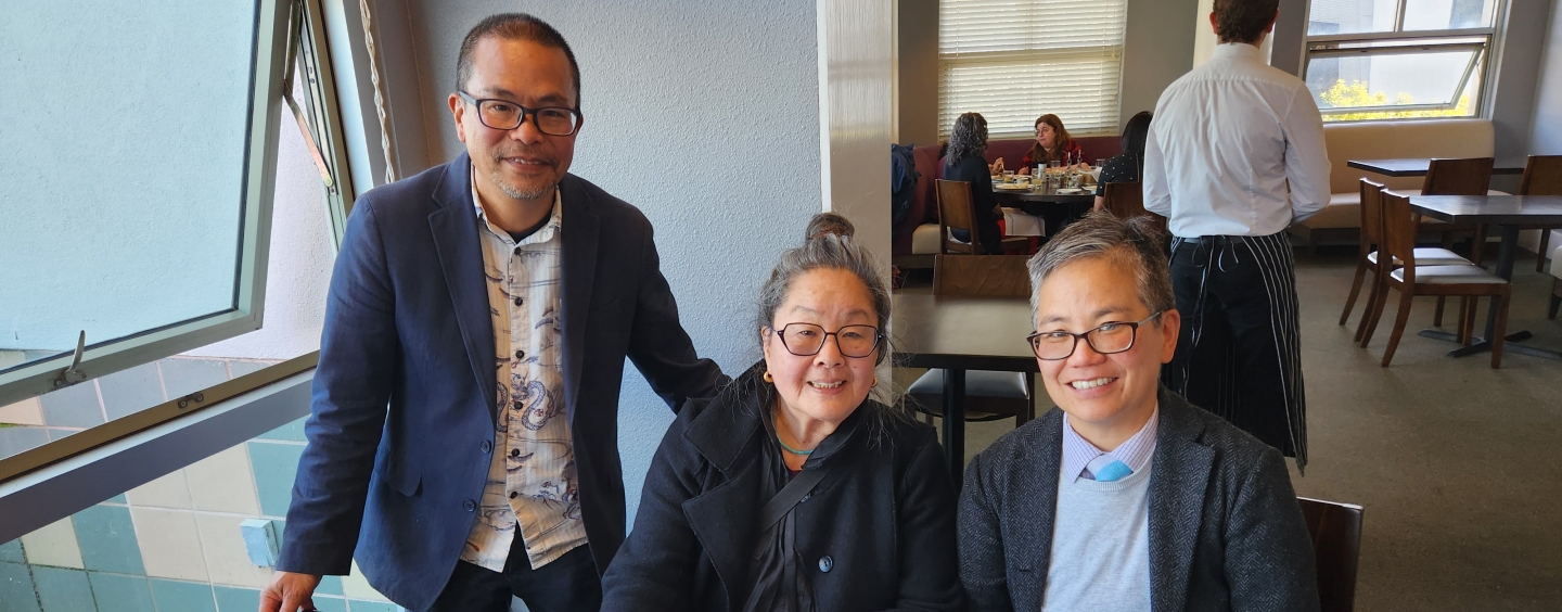 Three people at a table smiling. The person in the middle is holding a pen to paper on the table.