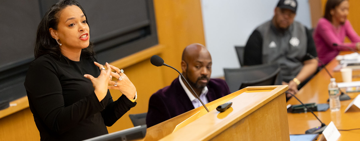 Dawn-Elissa Fischer uses hand gestures while speaking at a lectern dressed in black while other people seated next to hear watch