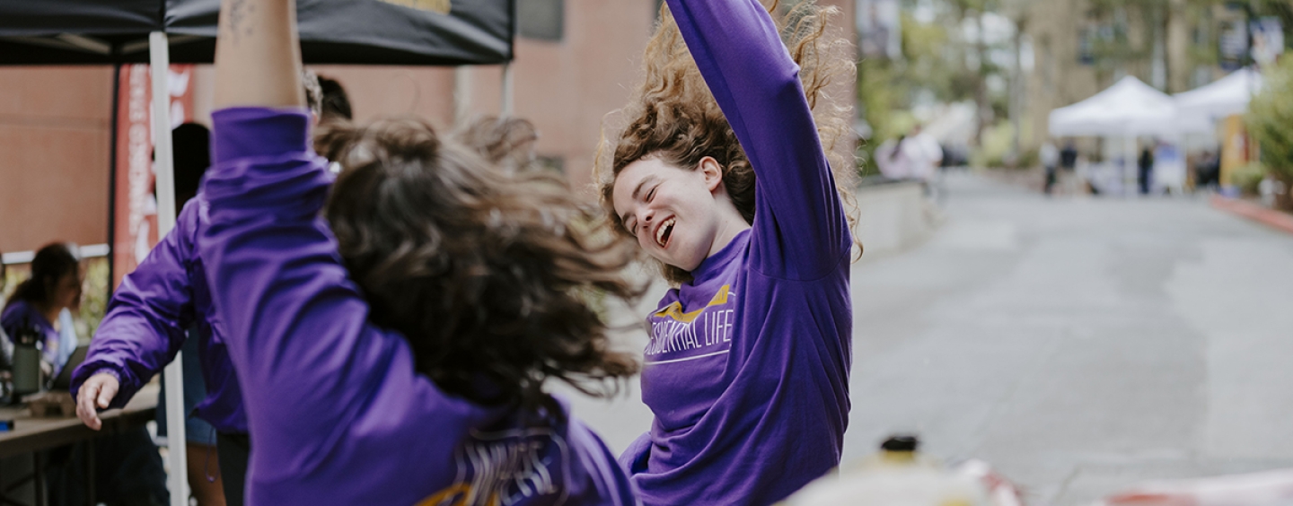 Two students standing outside raise their arms as they dance and cheer