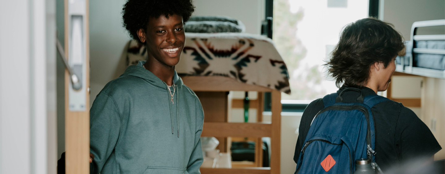 A student smiles while holding the door open to his room in the West Grove Commons while another person wearing a backpack walks into the room two with loft beds and a window visible behind them