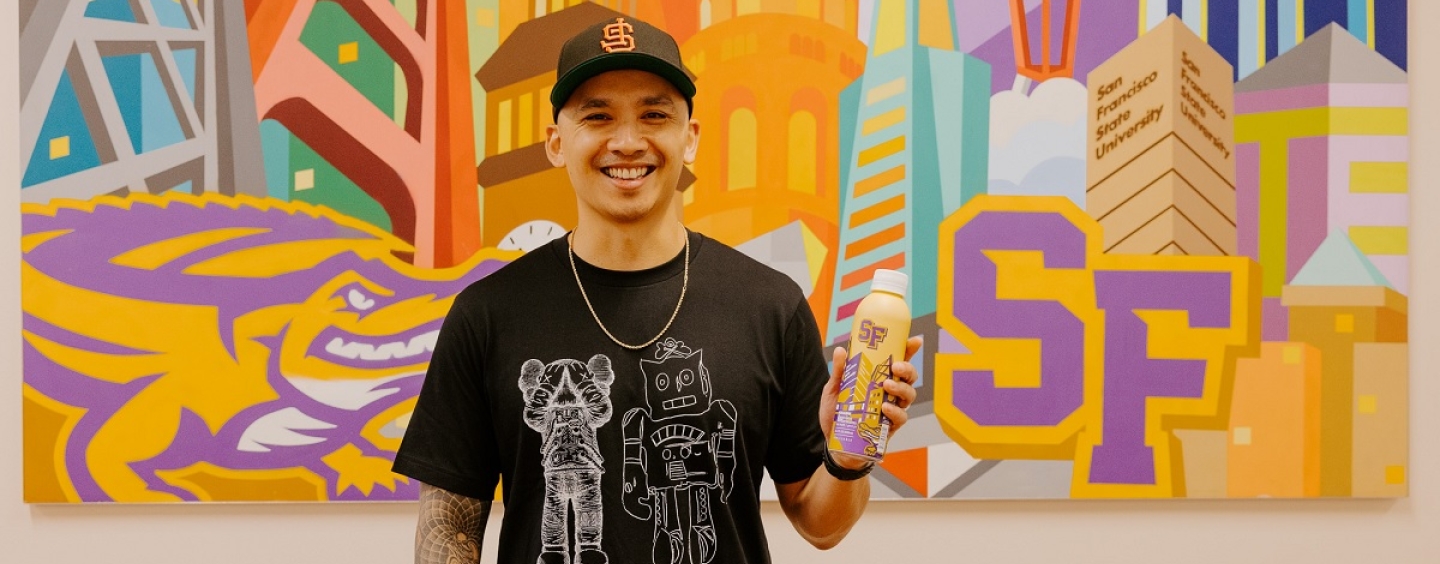 Jordan Herren stands in from of painting that he made that hands in SF State's administration building while holding a water bottle he illustrated