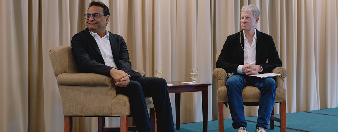 Laxman Narasimhan and Chris Larsen look to their right while seated on a stage between a table and in front of a curtain