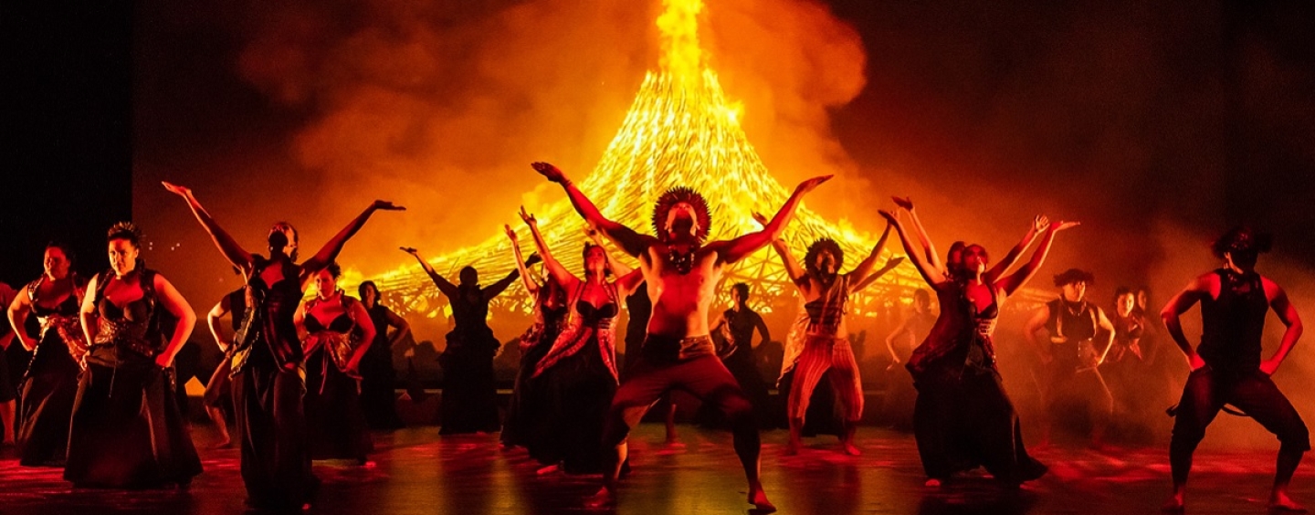 Dancers in traditional Hawaiian dress pose in front of a large fire