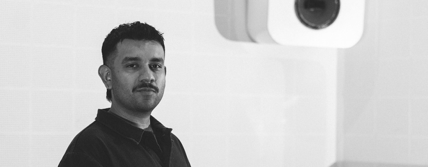 A black and white photo of Zach Pereyra standing in a studio with a speaker attached to the wall behind him