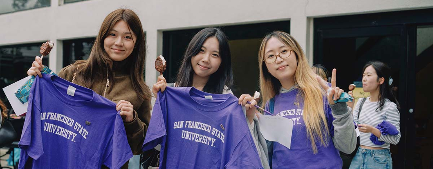 Three people hold up purple San Francisco State University T-shirts while standing and eating ice cream