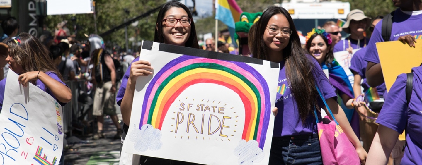 Two students marching in a parade stand in front of a hand-made sign saying "SF State Pride"