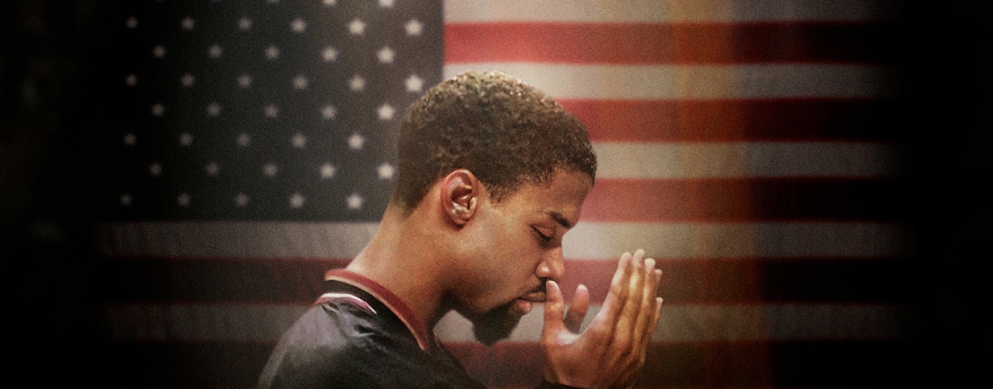 Mahmood Abdul-Rauf holds his hands in prayer in front of the USA flag while wearing a Denver Nuggets warmup jacket