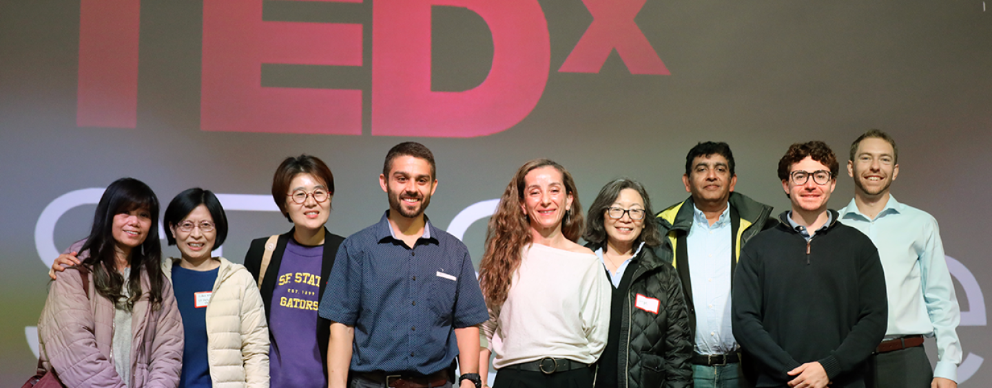 Nine students, faculty and administrators stand on stage in front of a screen projecting the TEDx logo