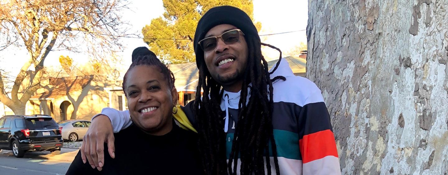 Marcus Angelo Bryant stands to the right of his mother, Theresa Thompson, with his right arm around her shoulders while both smile and stand next to a tree on a sunny day