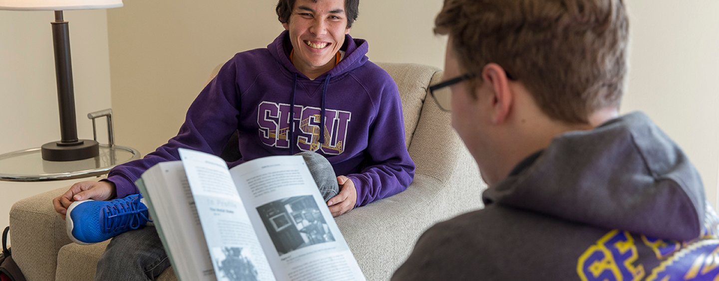 A person seated looks at a book while another person seated across from them smiles while wearing a purple SFSU hoodie