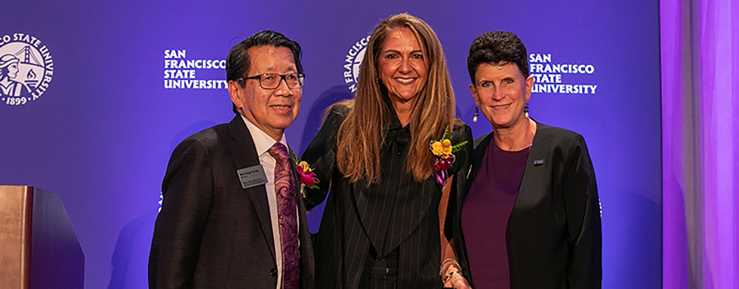 Three dressed up people pose together in front of a purple backdrop that says SFSU. 