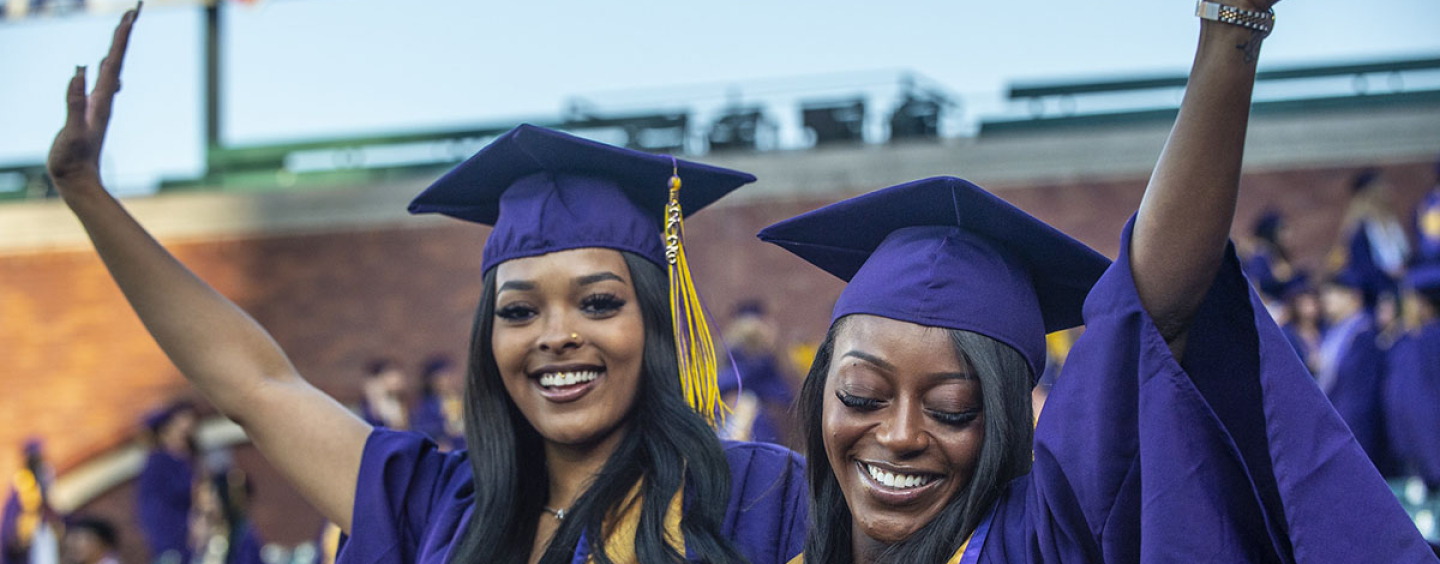 two people with long hair dressed in purple and gold reglia raise their arms in celebration