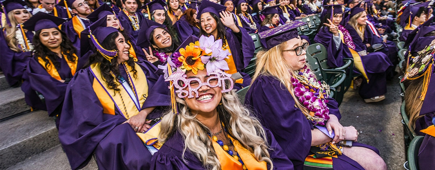 A graduate wears funny glasses that say while while sitting in a crowd of grads at Oracle Park 