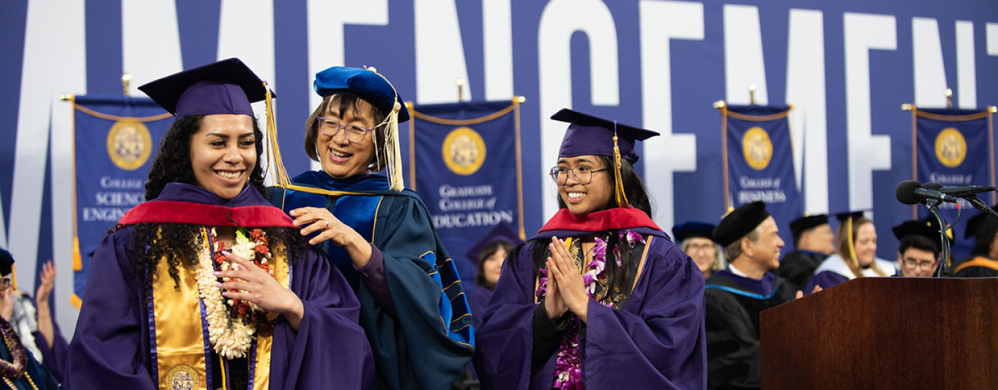 A student dressed in regalia gets hooded by a dean as another students watched proudly 