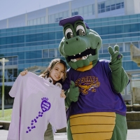 Student holding a lavendar Gator Giving Day shirt poses next to Alli Gator