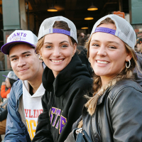 Three people smile while seated and wearing co-branded SFSU-San Francisco Giants baseball caps at Oracle Park
