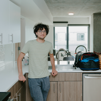 A student leans against the counter in his kitchenette in a residence hall with bags, a bicycle and a window behind him