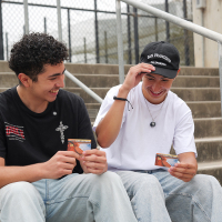 Two students smile and hold their Electronic Bank Transfer cards while seated next to each other on stairs outdoors on an overcast day