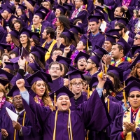 Several students stand in a stadium dressed in their caps and gown while one student smiles directly at the camera gesturing triumphantly