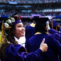 Student dressed in regalia in a ballpark gives the thumbs up 