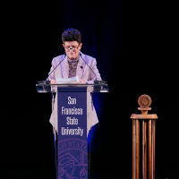 President Lynn Mahoney speaks behind a lectern decorated in purple with the text San Francisco State University in front of a black background and next to a wooden gavel 