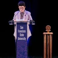 President Lynn Mahoney speaks behind a lectern decorated in purple with the text San Francisco State University in front of a black background and next to a wooden gavel 