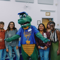 Seven people smile while posing for a photo with the Alli Gator mascot in the middle pointing their fingers horizontally and with one of the other people flashing a peace sign