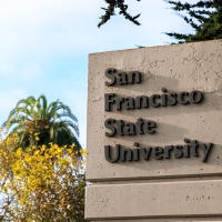 The San Francisco State University pylon sign surrounded by trees and next to a stoplight and a bicyclist road sign on a mostly sunny day
