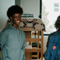 A student smiles while holding the door open to his room in the West Grove Commons while another person wearing a backpack walks into the room two with loft beds and a window visible behind them