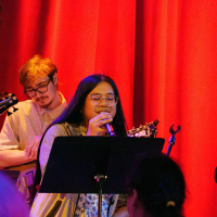 Anthony Joerel Garcia sings while holding a microphone and standing behind an easel and in front of an instrumentalist and a red curtain