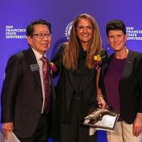 Three dressed up people pose together in front of a purple backdrop that says SFSU. 