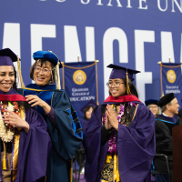 A student dressed in regalia gets hooded by a dean as another students watched proudly 